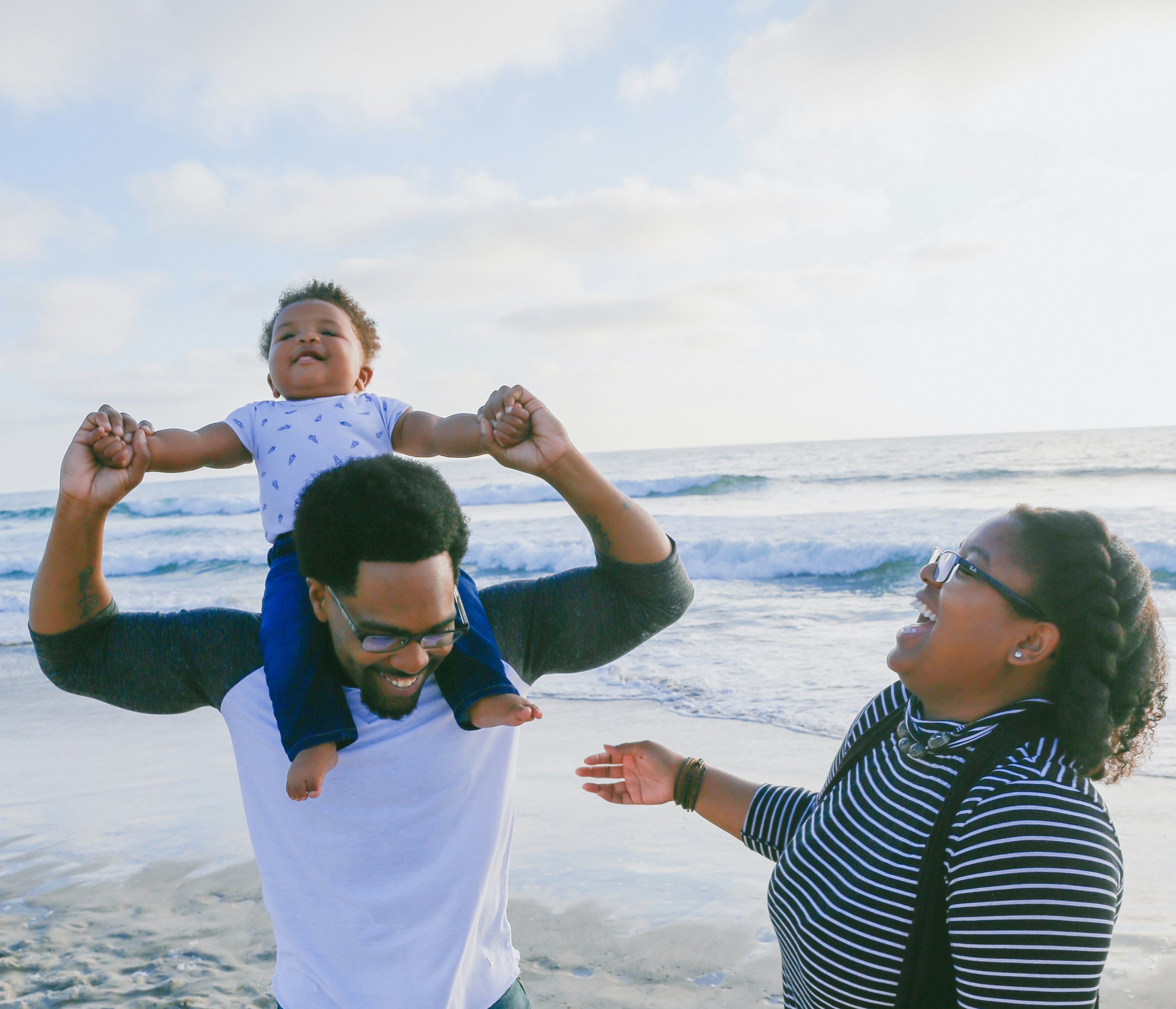 A father lifting his laughing baby above his head on the beach while the mother laughs beside them